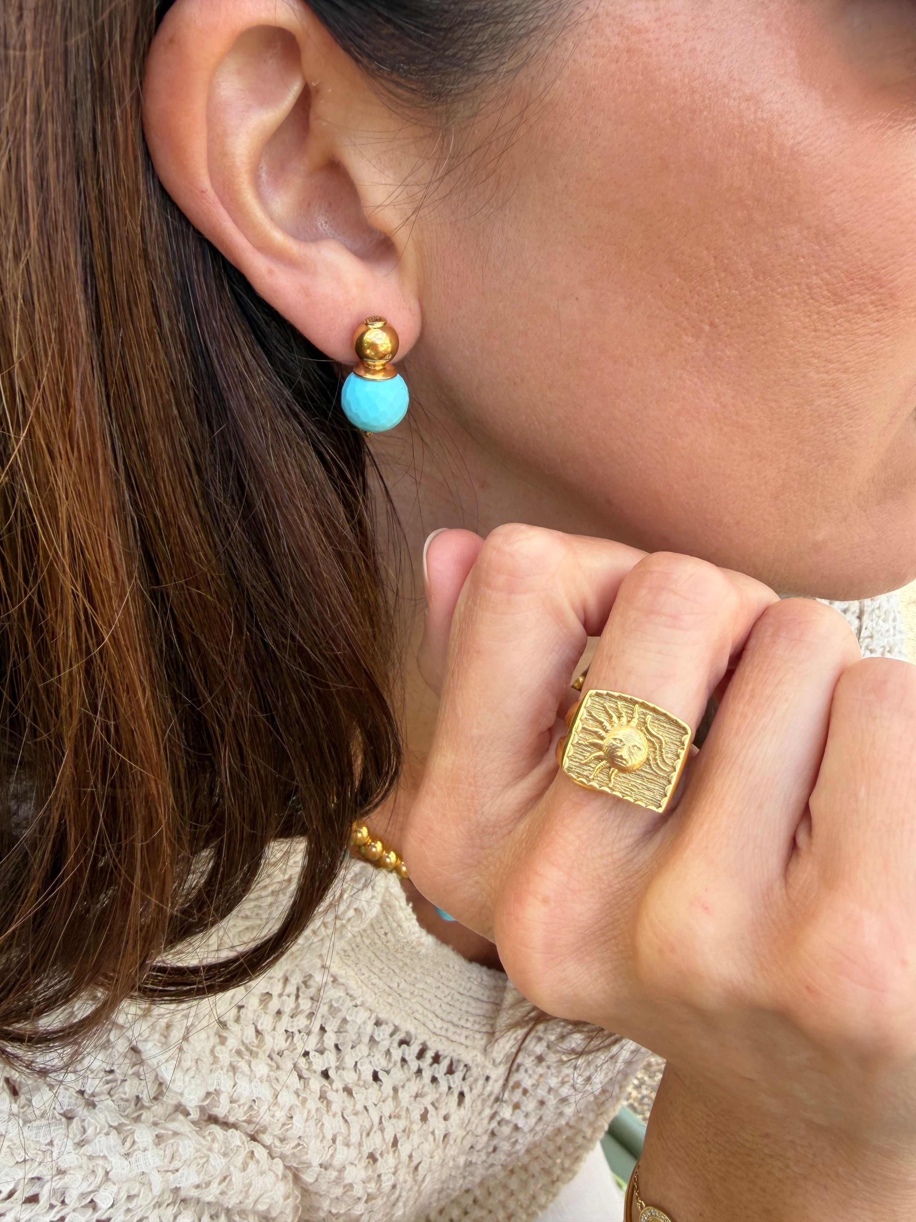 Close-up of a person wearing gold and turquoise earrings and a gold ring with a textured background.