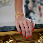 A close-up of a person's hand resting on an ornate gold and green marble table, wearing a delicate gold band ring with a small central stone and stacked gold bracelets.
