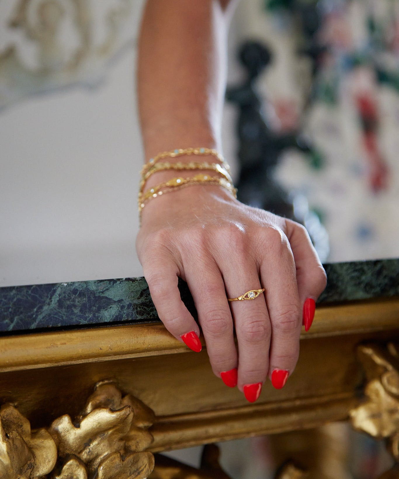 A close-up of a person's hand resting on an ornate gold and green marble table, wearing a delicate gold band ring with a small central stone and stacked gold bracelets.