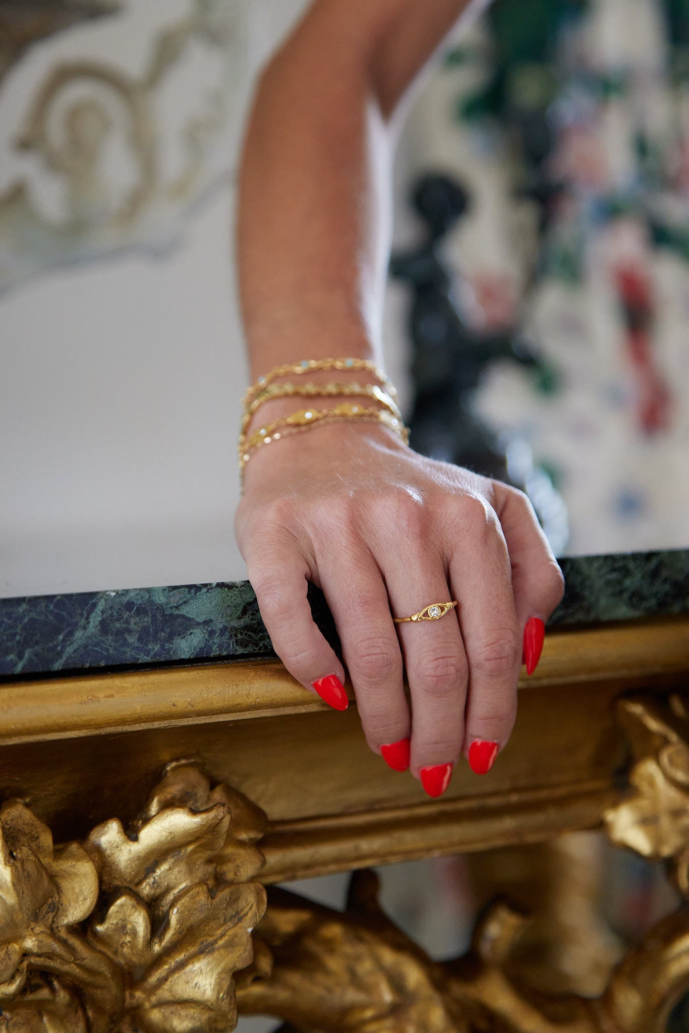 A close-up of a person's hand resting on an ornate gold and green marble table, wearing a delicate gold band ring with a small central stone and stacked gold bracelets.