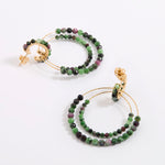 A pair of delicate gold double-hoop earrings, adorned with multiple strands of small, faceted beads in rich shades of green, dark charcoal, and purple, displayed against a white background.