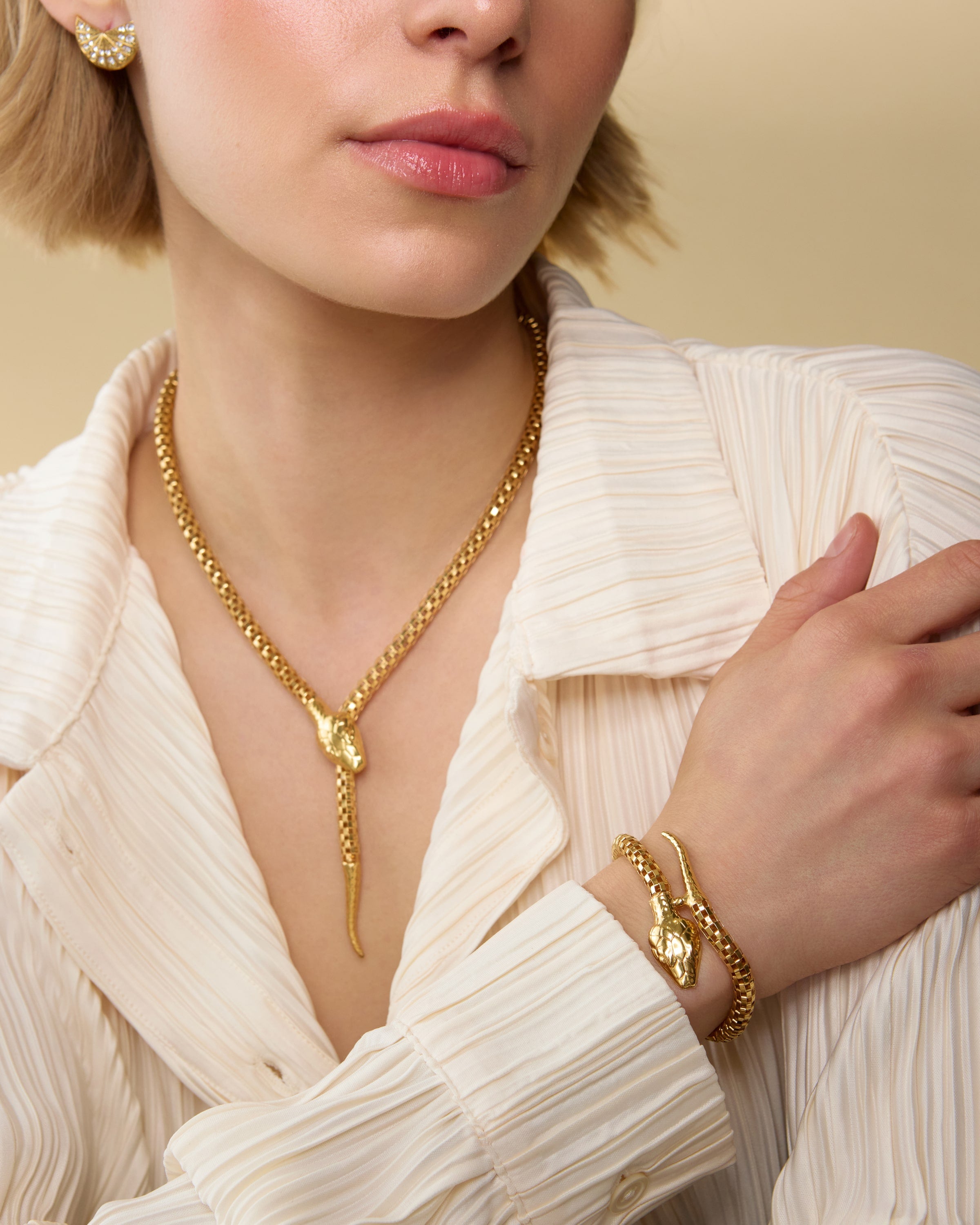 Model wearing gold snake motif bracelet on one wrist and necklace around her neck, with a cream shirt and gold fan stud earrings.