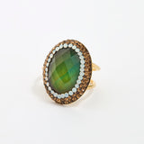 An oval ring featuring a faceted green gemstone, surrounded by a double-layered border of gold and opal stones, set on a polished gold band against a clean, plain white background.