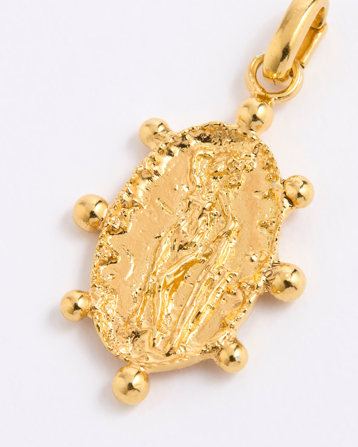 A close-up of a textured gold charm pendant featuring an embossed figure, surrounded by small gold beads, on a plain white background.