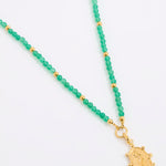 A green beaded necklace featuring small green gemstone beads separated by tiny gold spacer beads, with a textured gold pendant at the end, displayed on a white background.