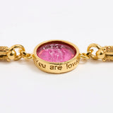 Gold link bracelet with round pendant in the centre with magenta glass and isciption 'you are loved' 