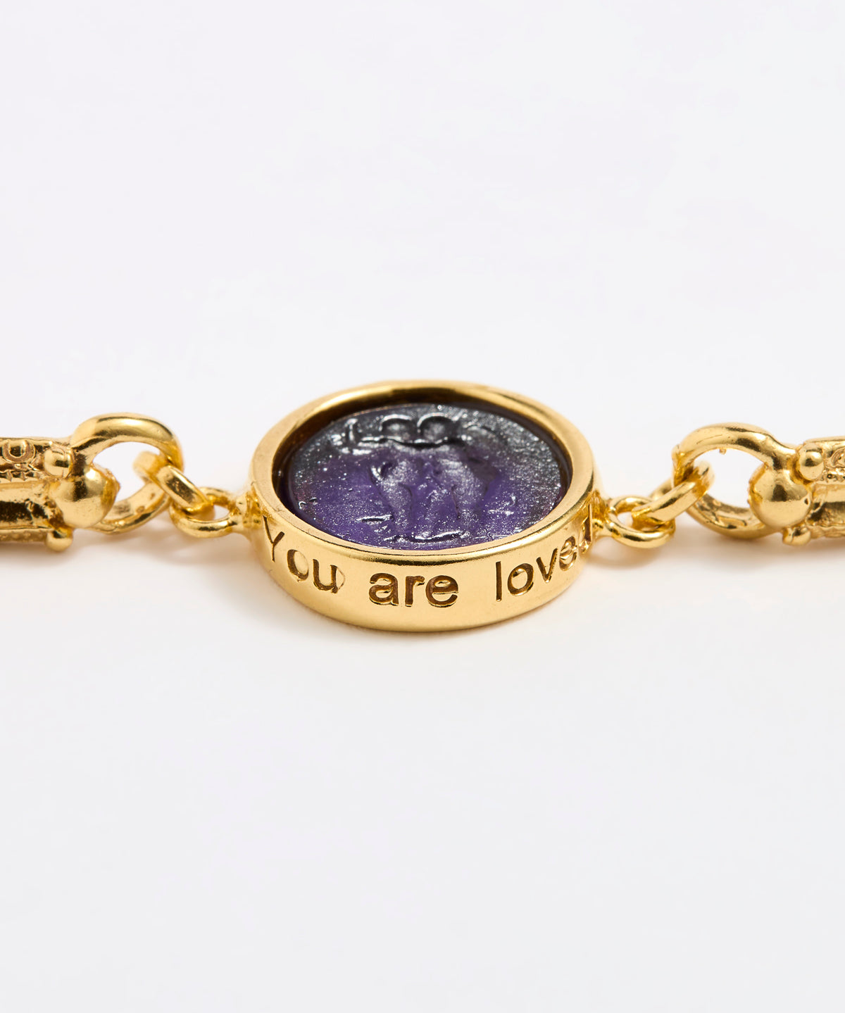 Close up of Gold link bracelet with round pendant in the centre with violet glass and inscription ' you are loved' 