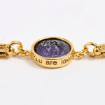 Close up of Gold link bracelet with round pendant in the centre with violet glass and inscription ' you are loved' 