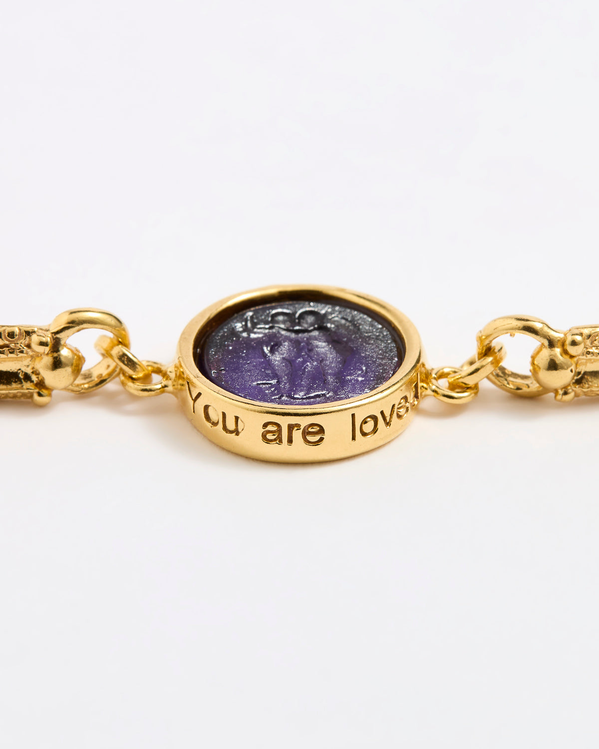 Close up of Gold link bracelet with round pendant in the centre with violet glass and inscription ' you are loved' 