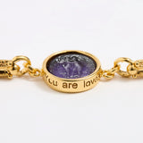 Close up of Gold link bracelet with round pendant in the centre with violet glass and inscription ' you are loved' 