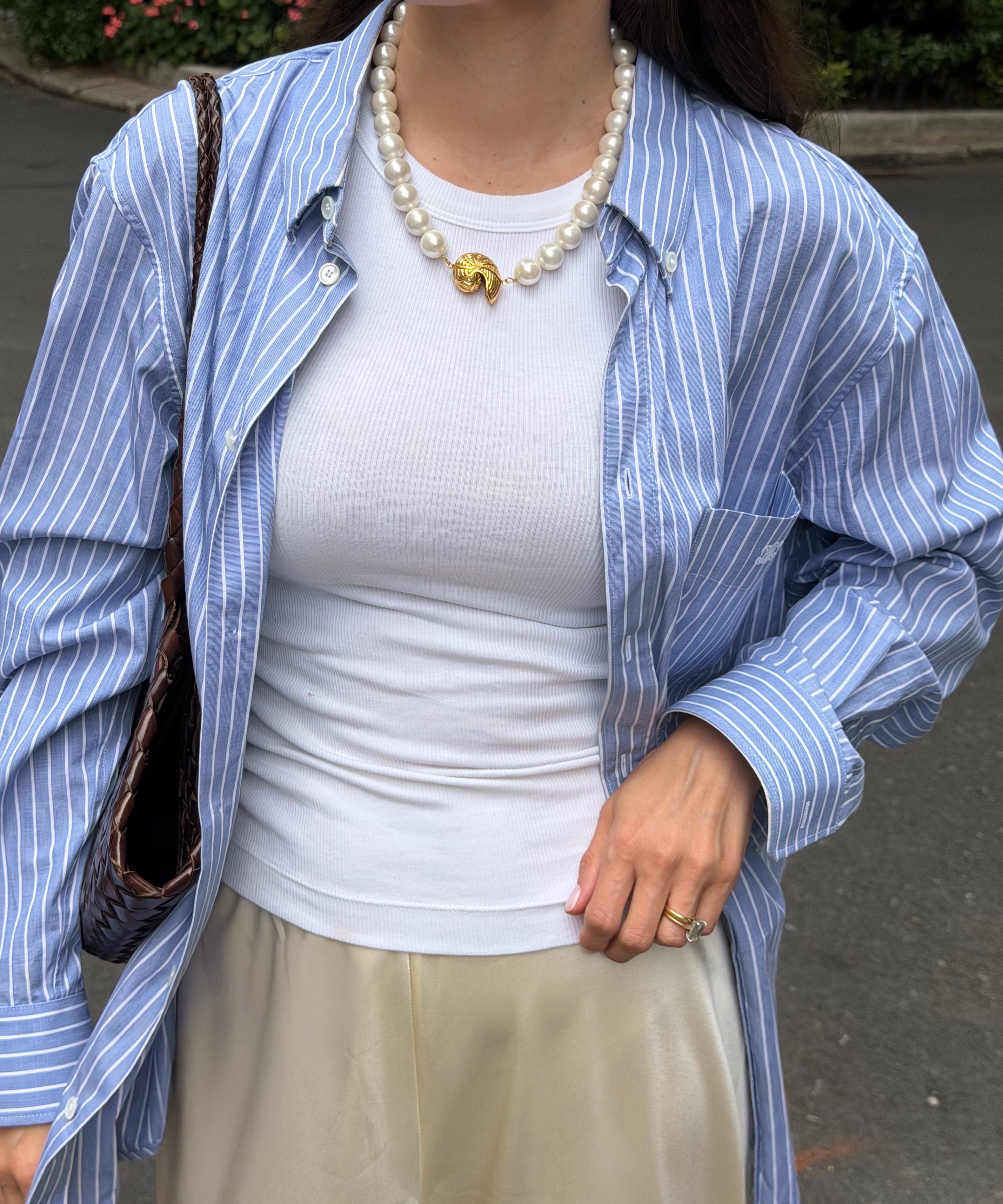 Person wearing a blue striped shirt over a white top with beige pants and a chunky pearl necklace,  holding a brown bag.