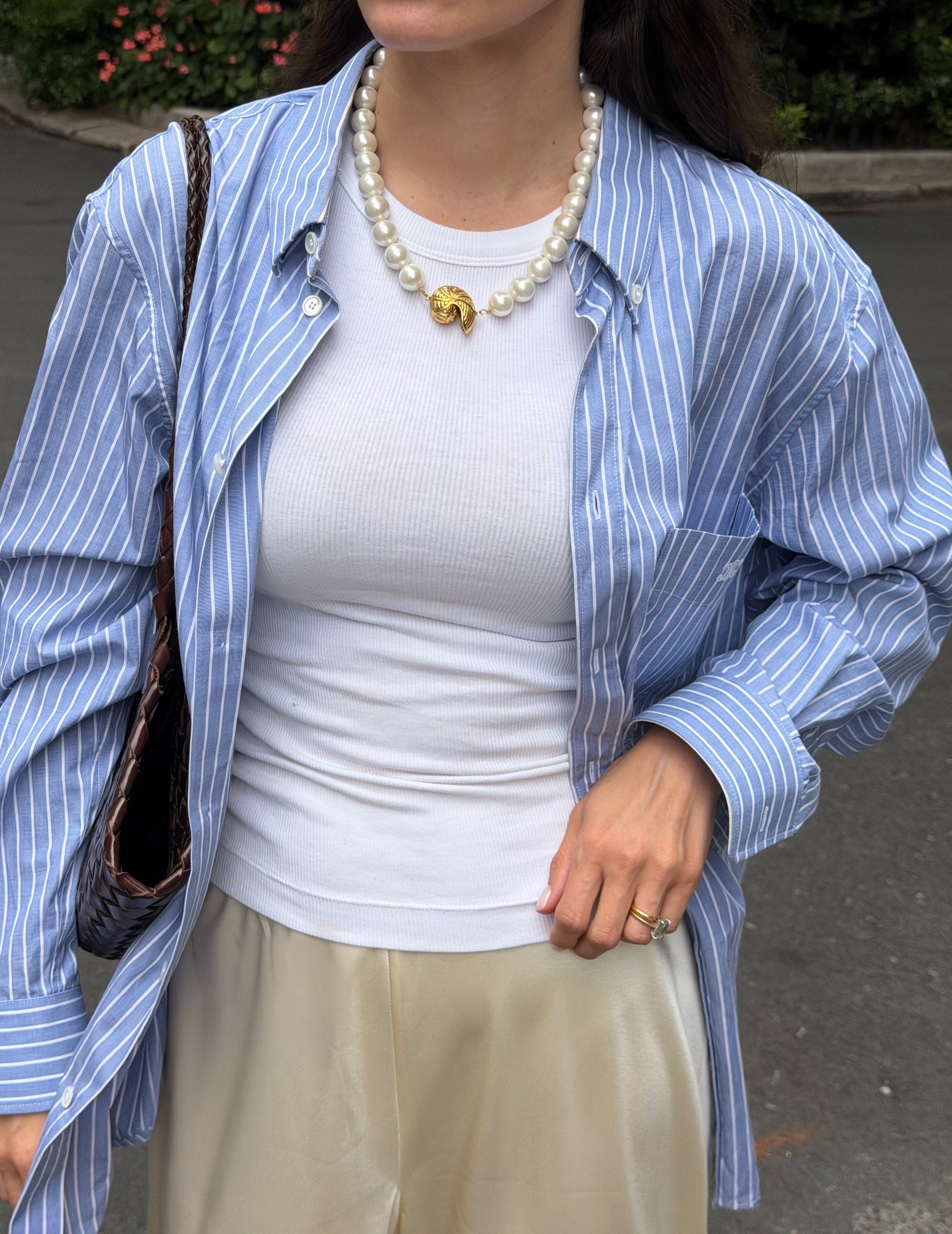 Person wearing a blue striped shirt over a white top with beige pants and a chunky pearl necklace,  holding a brown bag.