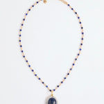 A beaded necklace featuring deep blue lapis lazuli stones and gold-toned spacer beads, with a large oval lapis lazuli pendant encircled by small white stones, displayed on a clean white background.