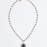 A beaded necklace featuring deep blue lapis lazuli stones and gold-toned spacer beads, with a large oval lapis lazuli pendant encircled by small white stones, displayed on a clean white background.