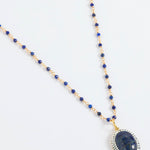 A beaded necklace featuring deep blue lapis lazuli stones and gold-toned spacer beads, with a large oval lapis lazuli pendant encircled by small white stones, displayed on a clean white background.