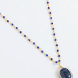 A beaded necklace featuring deep blue lapis lazuli stones and gold-toned spacer beads, with a large oval lapis lazuli pendant encircled by small white stones, displayed on a clean white background.