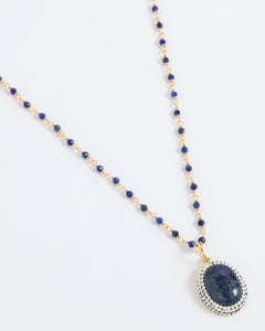A beaded necklace featuring deep blue lapis lazuli stones and gold-toned spacer beads, with a large oval lapis lazuli pendant encircled by small white stones, displayed on a clean white background.