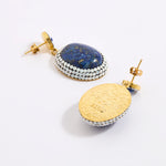 A pair of gold and lapis lazuli earrings shown on a white background, with one earring facing forward to display the blue lapis lazuli stone and white opal crystal border, and the other showing the textured gold back with a post.