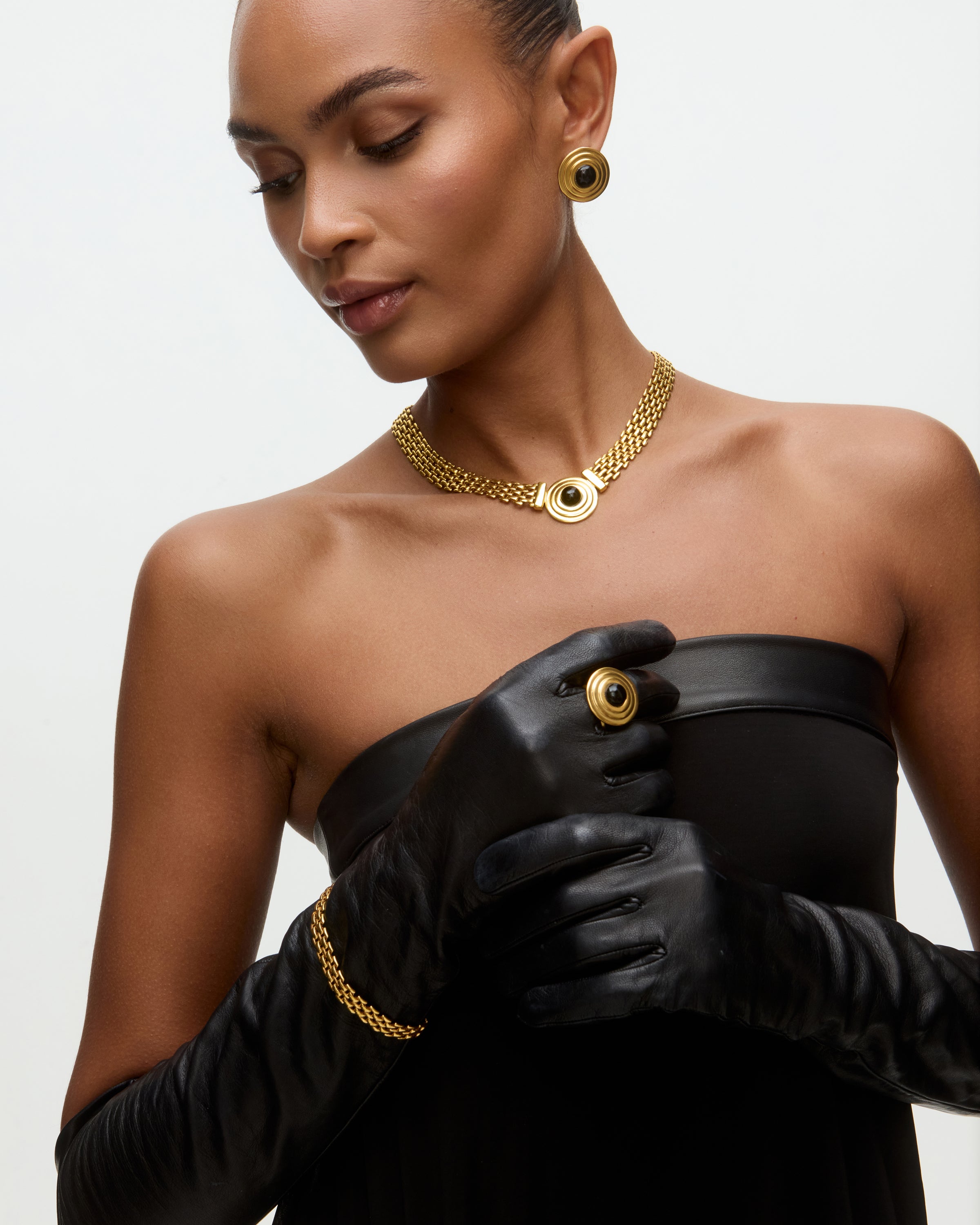 Image of a lady wearing black dress, gold chain bracelet, matching choker with round onyx detail and matching stud earrings.