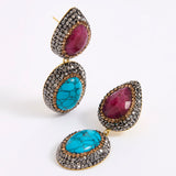 A pair of gold-tone drop earrings featuring oval turquoise and teardrop ruby gemstones, surrounded by a dark silver crystal setting.