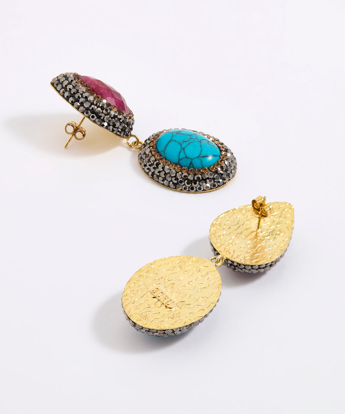 A pair of gold-toned statement earrings displayed against a white background. One earring features a vibrant turquoise stone, surrounded by dark silver crystals and accented with a ruby teardrop gemstone above. The other earring is shown from the back, highlighting its textured gold finish and dark silver crystal border.