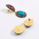 A pair of gold-toned statement earrings displayed against a white background. One earring features a vibrant turquoise stone, surrounded by dark silver crystals and accented with a ruby teardrop gemstone above. The other earring is shown from the back, highlighting its textured gold finish and dark silver crystal border.
