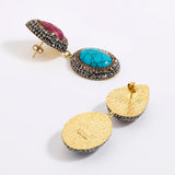 A pair of gold-toned statement earrings displayed against a white background. One earring features a vibrant turquoise stone, surrounded by dark silver crystals and accented with a ruby teardrop gemstone above. The other earring is shown from the back, highlighting its textured gold finish and dark silver crystal border.