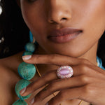 Model shot wearing chunky ring with pink centre stone and surrounded with crystals with hand under chin.  Model also wearing a delicate gold evil eye necklace
