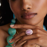 Model shot wearing chunky ring with pink centre stone and surrounded with crystals with hand under chin.  Model also wearing a delicate gold evil eye necklace