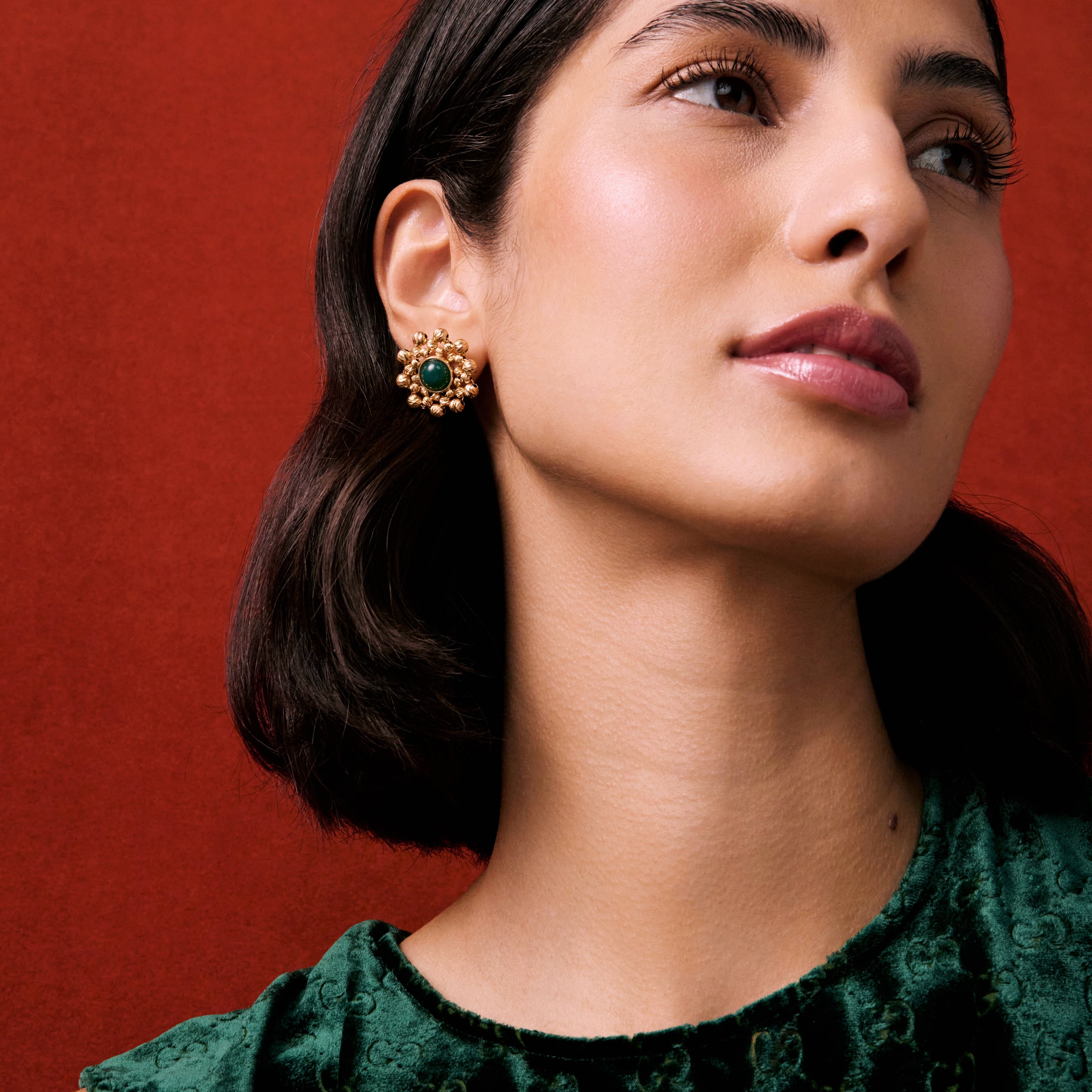 Product shot of lady wearing flower shaped beaded earrings featuring a central dark green gemstone on a red background