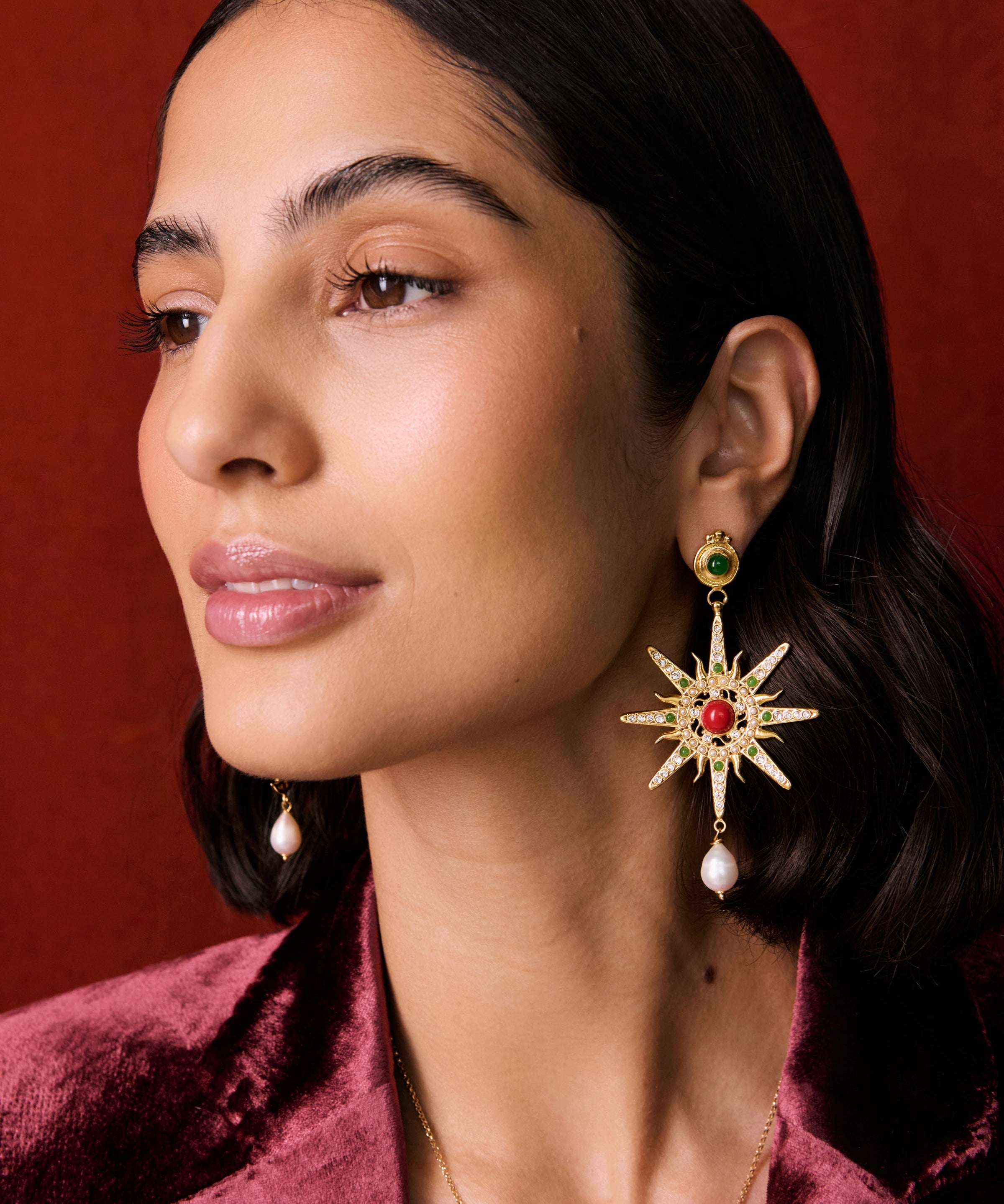Model shot of lady wearing a mis-matched earring, one featuring a star. Detailed with hanging pearls and colourful green and red stones, on a red background.
