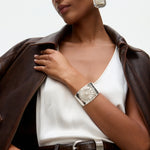 Woman wearing a brown leather jacket, white top, and silver watch with a neutral background and statement silver earrings and cuff