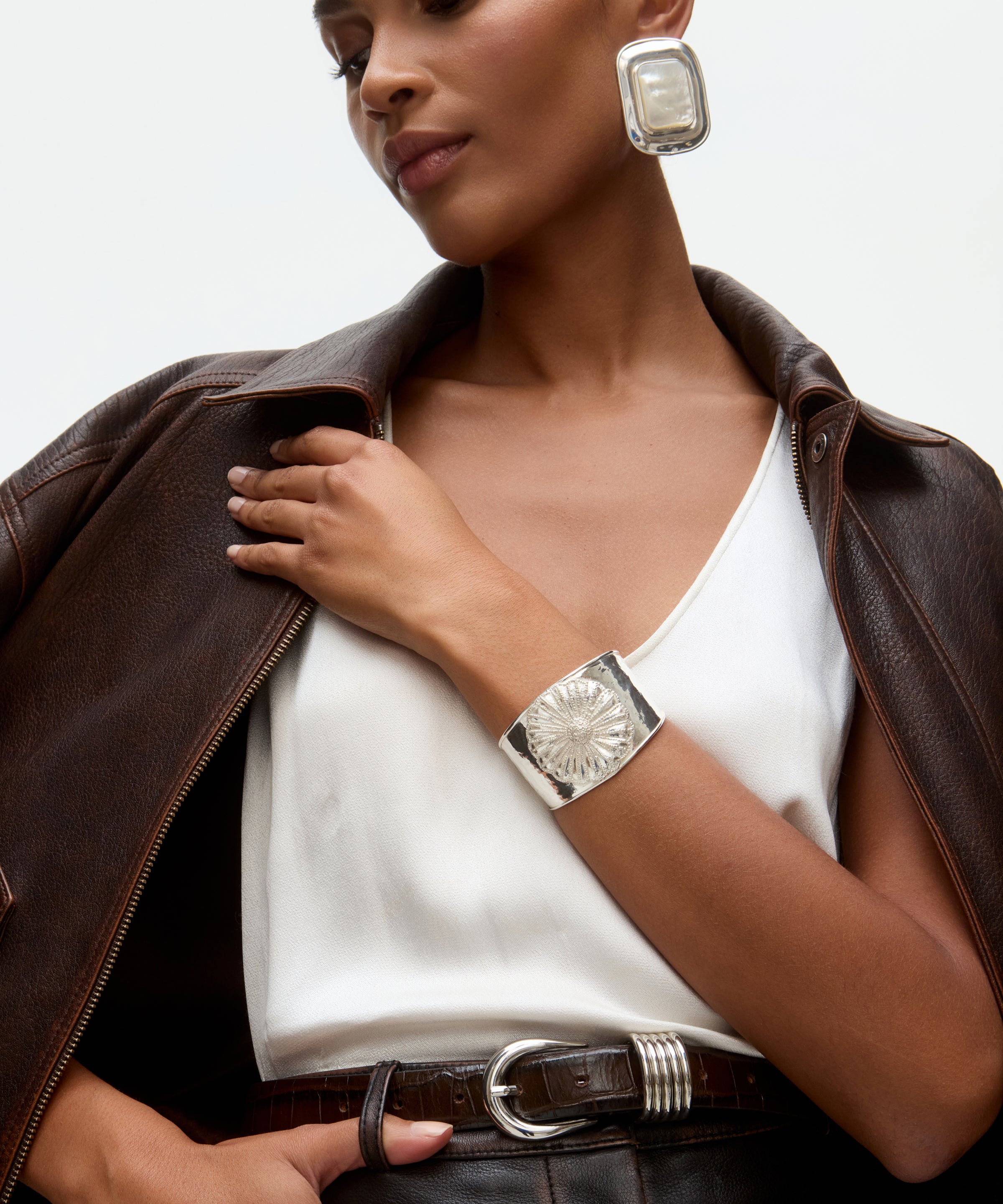 Woman wearing a brown leather jacket, white top, and silver watch with a neutral background and statement silver earrings and cuff