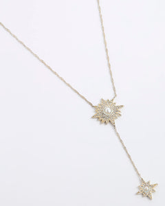 A gold-toned Y-necklace featuring two starburst pendants, each adorned with clear crystals and a central white pearl, displayed on a clean white background.