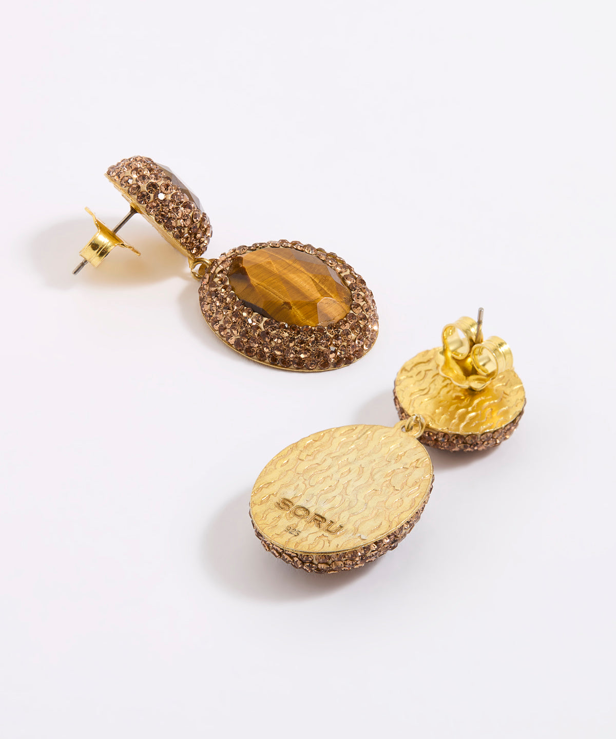 A pair of gold-toned dangle earrings with textured pavé settings, featuring one oval stone with a tiger's eye pattern and one flat textured gold disc, shown on a white background.