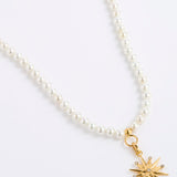 A white pearl necklace featuring a gold 12-pointed star charm pendant, embellished with sparkling clear crystals, presented on a white background.