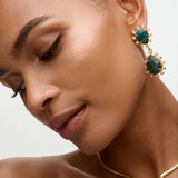 Close-up of a woman wearing gold earrings and necklace with green stones on a neutral background