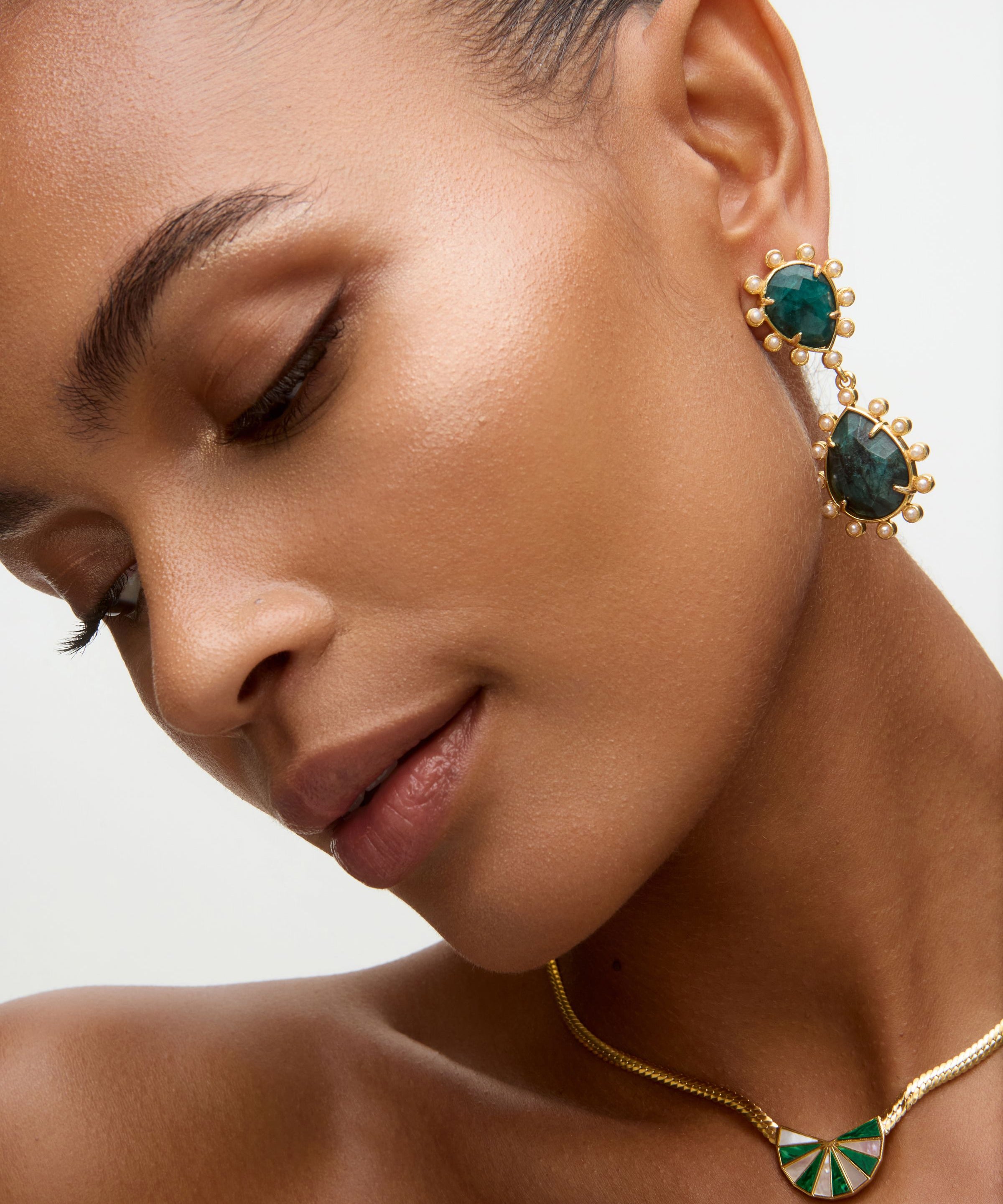 Close-up of a woman wearing gold earrings and necklace with green stones on a neutral background