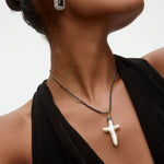 Close-up of a woman wearing an onyx and crystal earring and a necklace with a white baroque pearl pendant in the shape of a cross on a neutral background