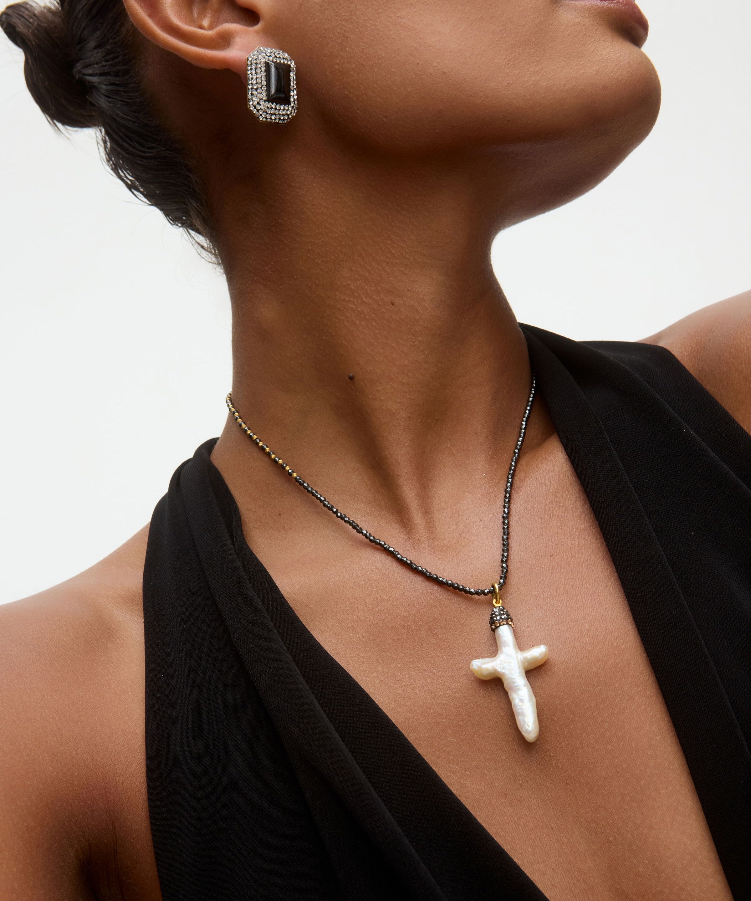 Close-up of a woman wearing an onyx and crystal earring and a necklace with a white baroque pearl pendant in the shape of a cross on a neutral background