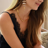 A woman wearing a gold geometric chain necklace and matching drop earrings with stacked white stone-like discs, shown in a close-up lifestyle shot.