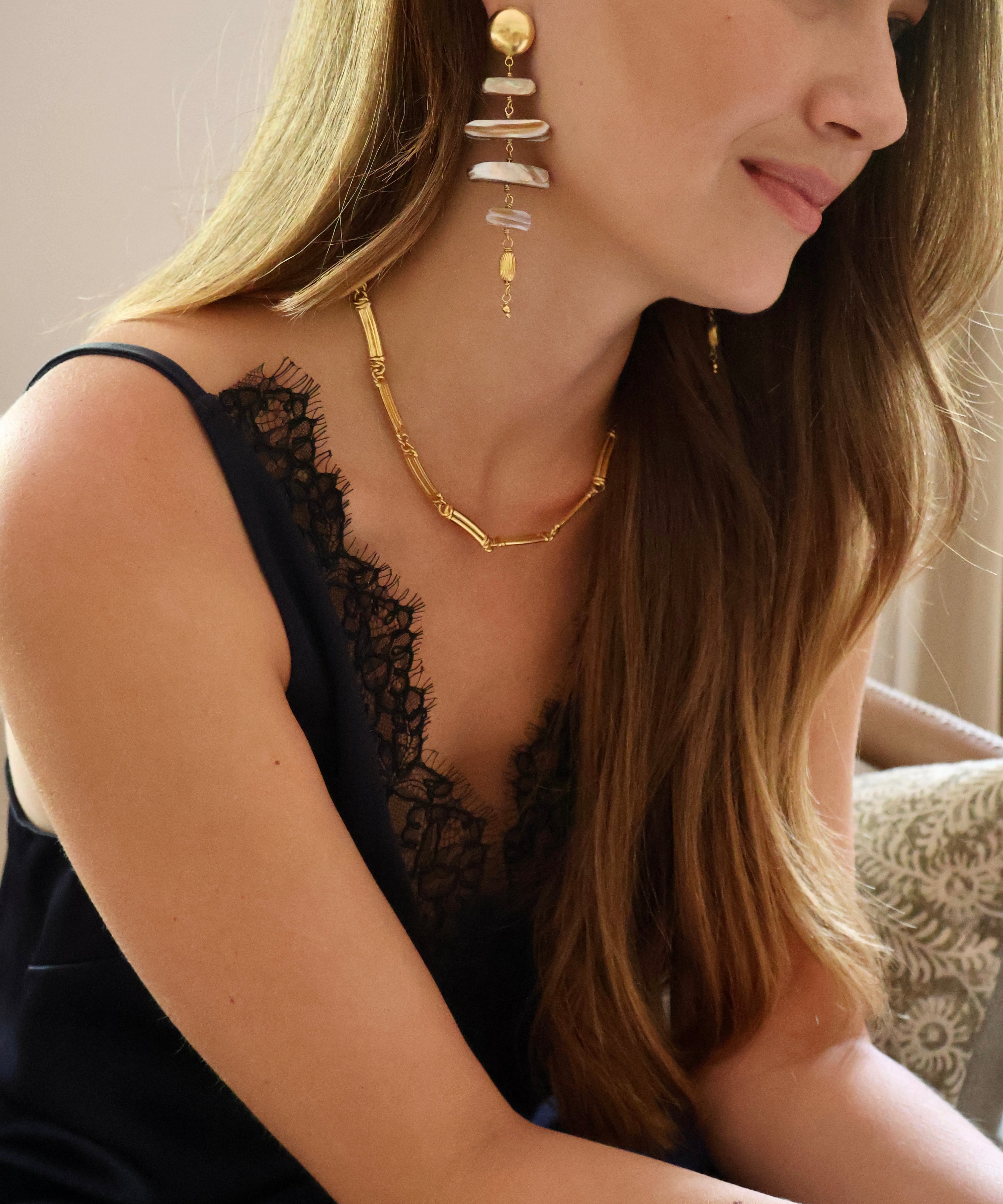 A woman wearing a gold geometric chain necklace and matching drop earrings with stacked white stone-like discs, shown in a close-up lifestyle shot.