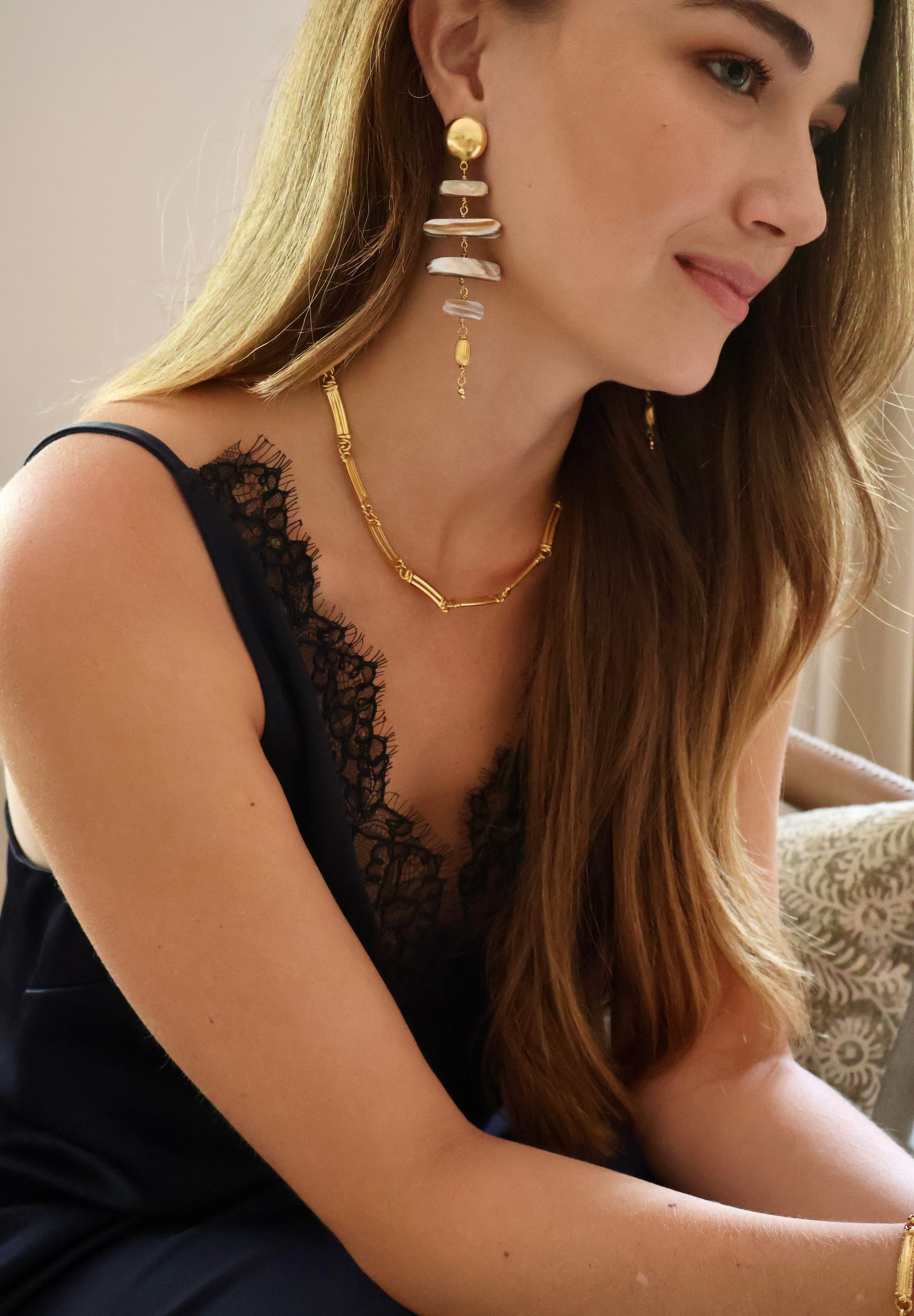 A woman wearing a gold geometric chain necklace and matching drop earrings with stacked white stone-like discs, shown in a close-up lifestyle shot.