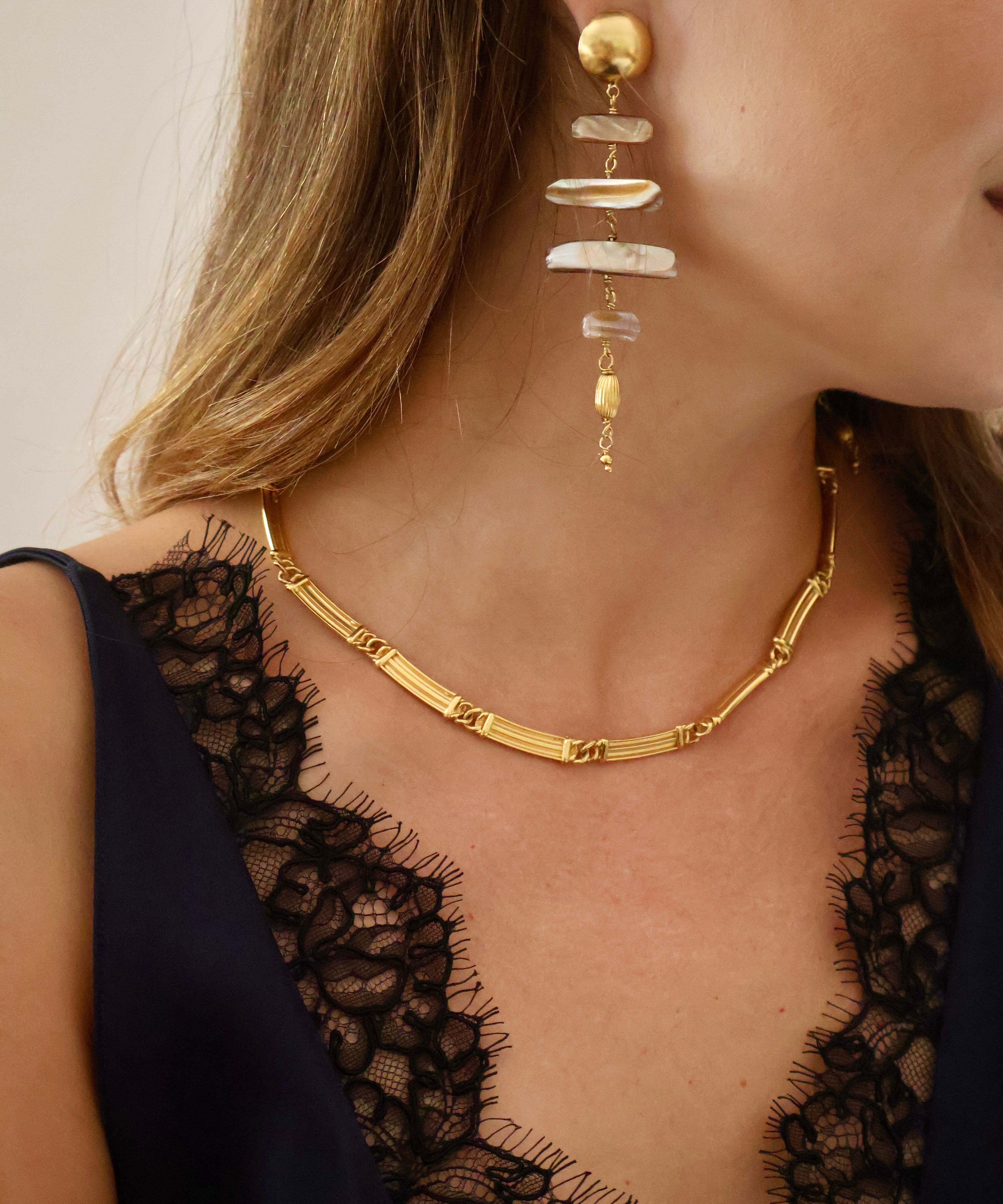 Close-up of a woman wearing a gold chain necklace and a matching gold drop earring with stacked shell-like white and beige elements, against a soft light background.