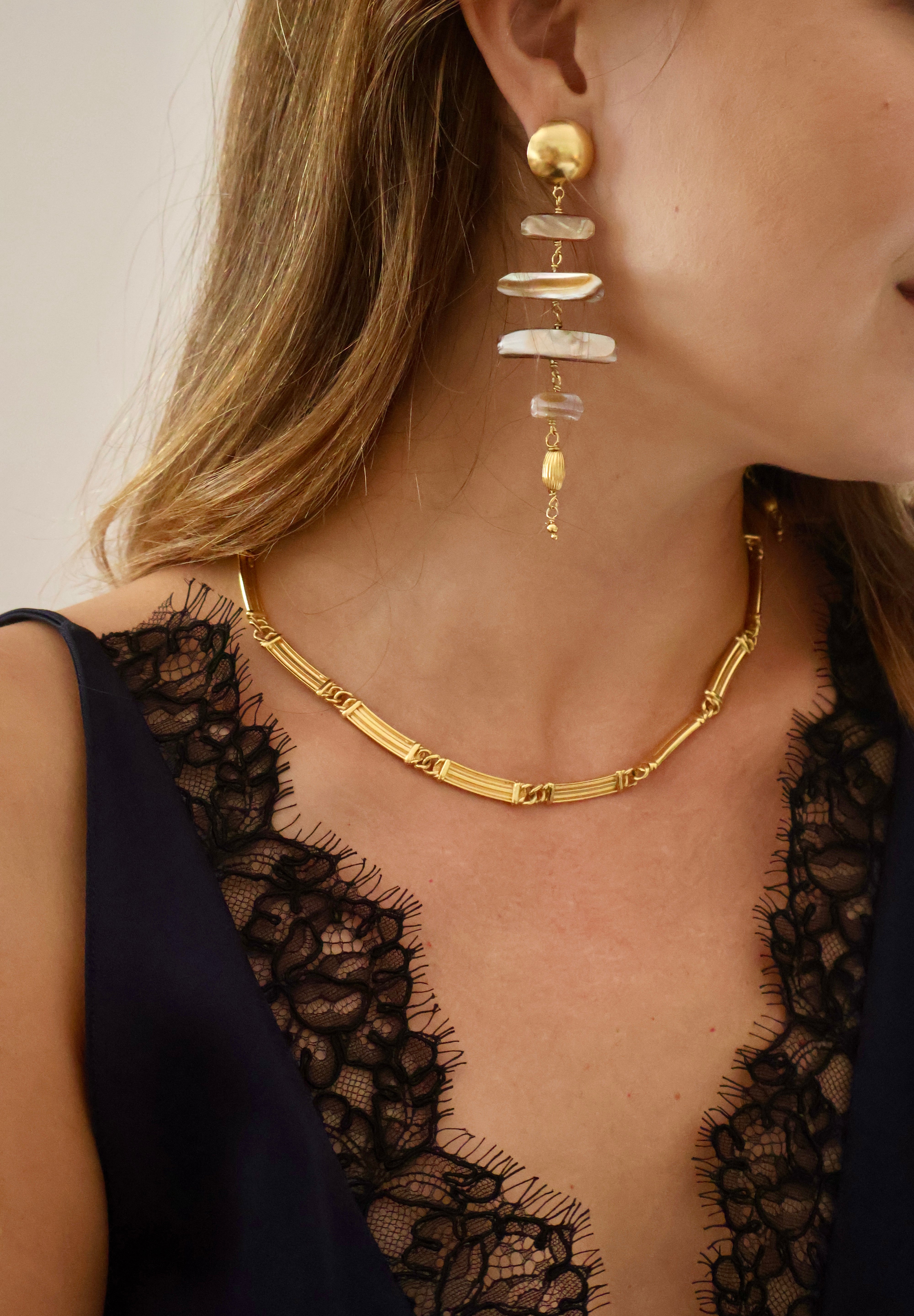 Close-up of a woman wearing a gold chain necklace and a matching gold drop earring with stacked shell-like white and beige elements, against a soft light background.