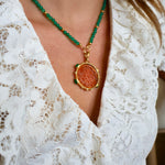 A close-up of a person wearing a necklace with green faceted beads, a gold-tone chain, and a round pendant featuring a textured orange-brown center surrounded by a gold-tone border. The person is wearing a white lace top.