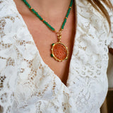 A close-up of a person wearing a necklace with green faceted beads, a gold-tone chain, and a round pendant featuring a textured orange-brown center surrounded by a gold-tone border. The person is wearing a white lace top.