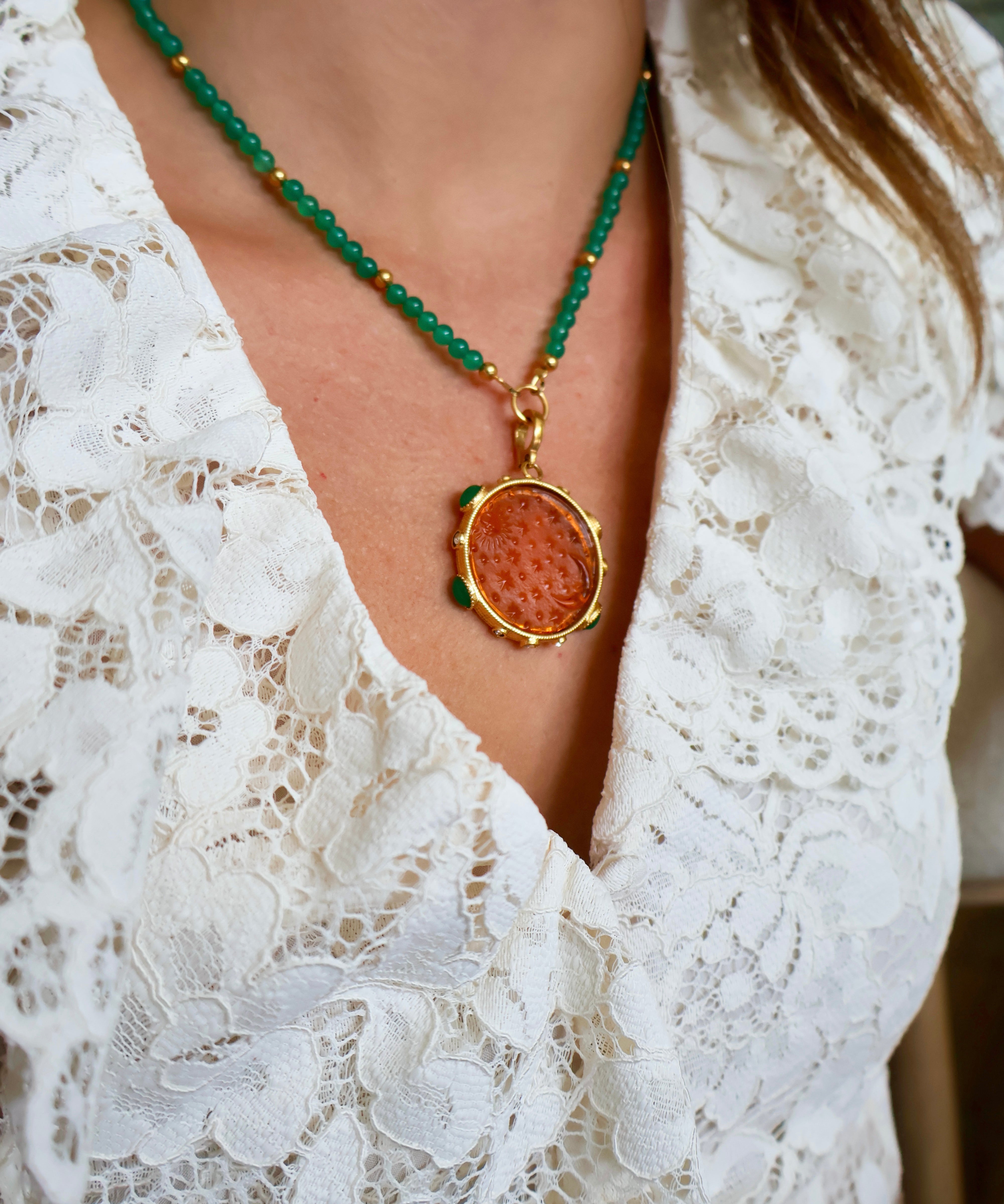 A close-up of a person wearing a necklace with green faceted beads, a gold-tone chain, and a round pendant featuring a textured orange-brown center surrounded by a gold-tone border. The person is wearing a white lace top.