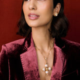 Model shot of lady wearing a gold cross pendant necklace and velvet blazer and gold ring. The necklace features pastel coloured agate gemstones, pearls and intricate filigree hung from a textured beaded chain.