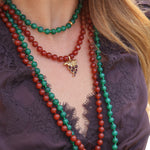 A woman wears two beaded necklaces, one red and the other green with a delicate grape charm. The necklaces are styled with a beautiful purple lace top for a bold and elegant look.